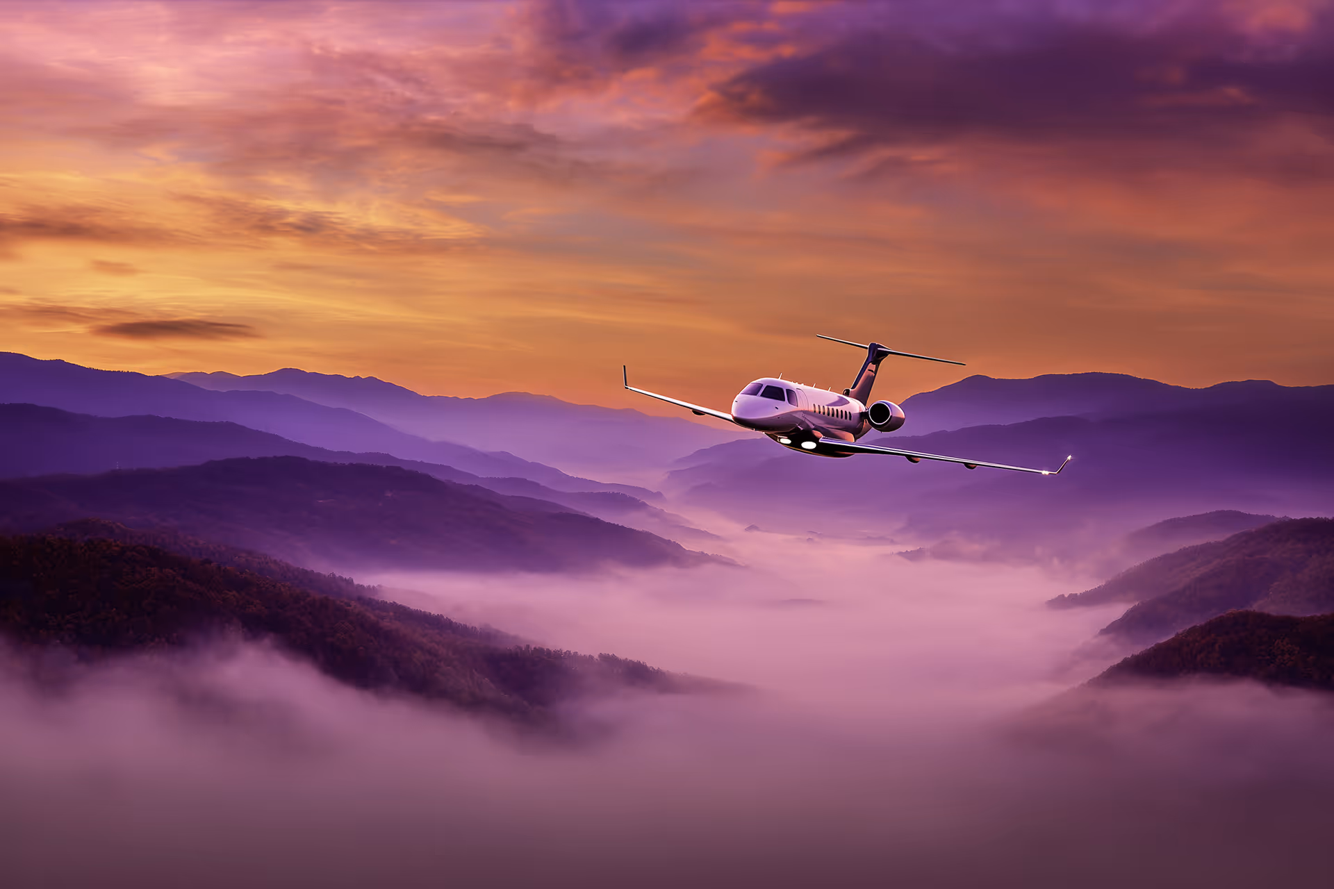 Private jet flying over misty mountains during a vibrant sunset with purple and orange sky.